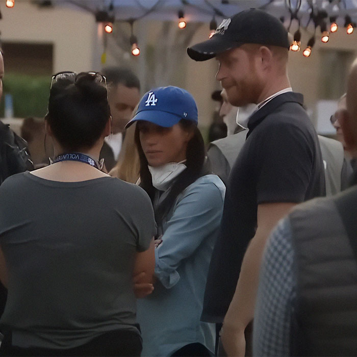 A woman in a blue cap stands with a man in a black cap, engaged in a conversation; Meghan Markle, Prince Harry related scene.