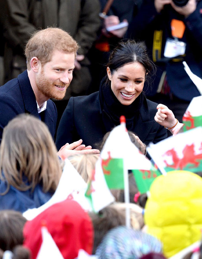 Royal couple engaging with children during a public event, amidst a sea of waving flags.