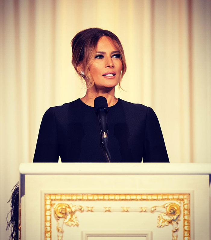 Melania Trump standing at a podium in a black dress, delivering a speech as First Lady.