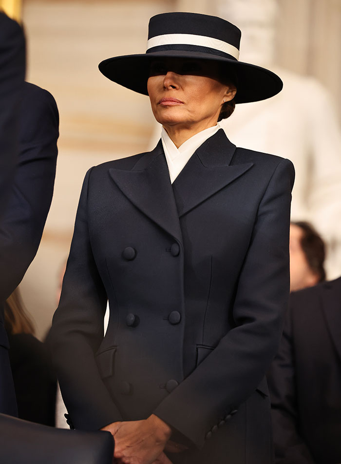 Melania Trump in a dark coat and hat, standing with a neutral expression during a public event.