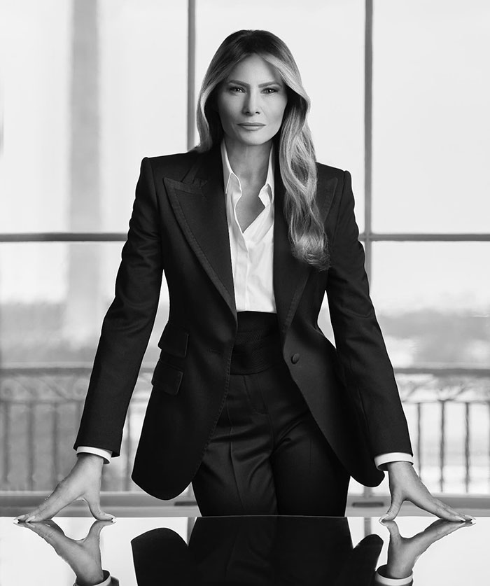 Melania Trump in a formal portrait, wearing a tailored black suit, standing confidently in a windowed room.