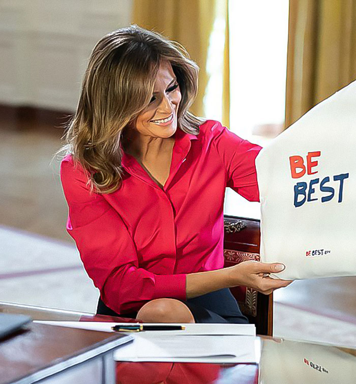 Melania Trump holding "Be Best" material, seated in an elegant room, wearing a bright pink blouse.