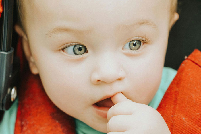 A curious baby with big eyes and a finger in its mouth, wearing a red outfit.