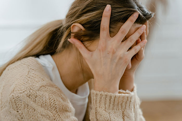 Person looking distressed, covering face with hands, wearing a beige sweater, sitting indoors.