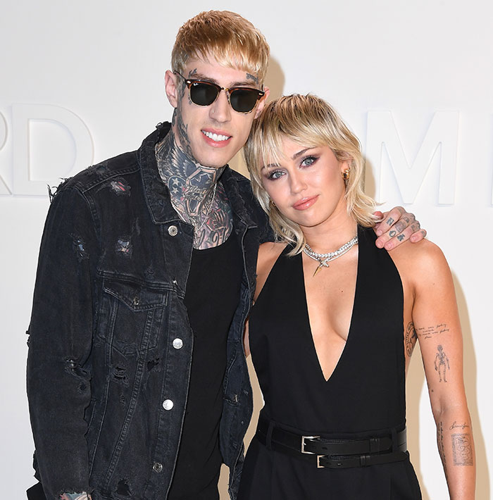 Two people posing together, one with tattoos and sunglasses, the other in a black dress; Cyrus family related.