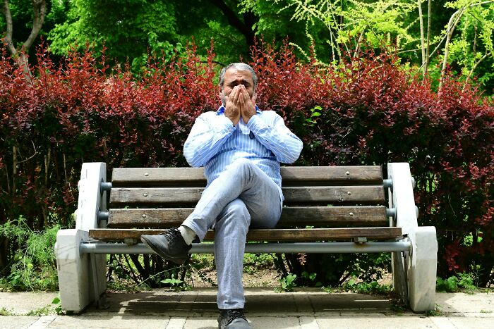 Man in a blue shirt covering face, sitting on a park bench, surrounded by greenery, symbolizing social norms.