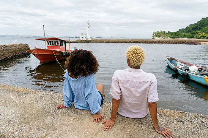 A man and woman sitting by the waterfront, boats in view, symbolizing a relationship and proposal moment in a crowd.