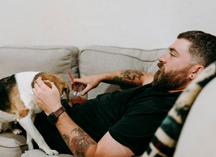 Man relaxing on a couch with a drink, petting a dog, showcasing entitled behavior.