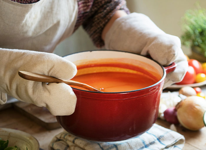 A red pot of spicy soup being stirred with love, held by a person wearing oven mitts.