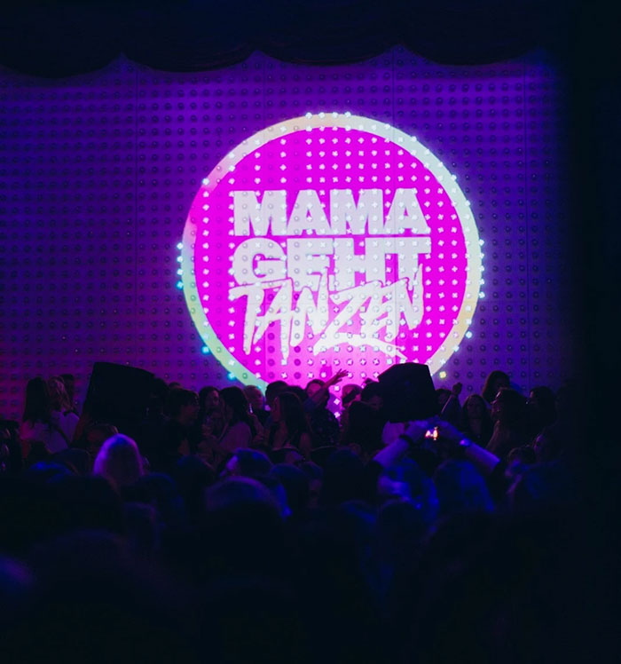 Nightclub scene with "Mama Geht Tanzen" projected on wall, redefining partying post-baby for moms.