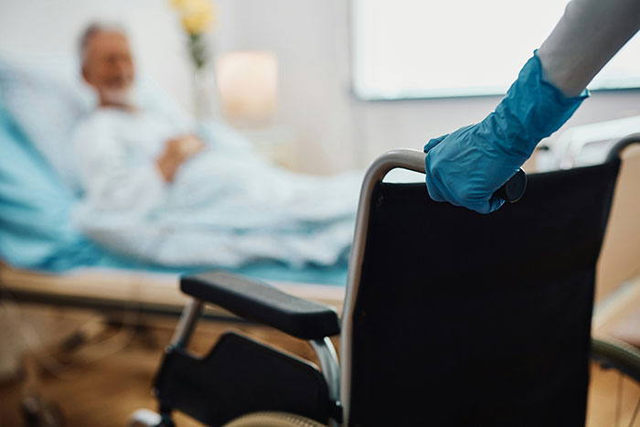 Gloved hand on wheelchair handle near hospital bed, conveying touching care moment in a healthcare setting.