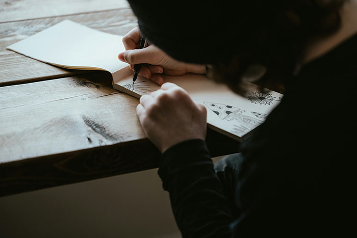 Person drawing in a sketchbook on a wooden table, focusing on capturing touching moments.