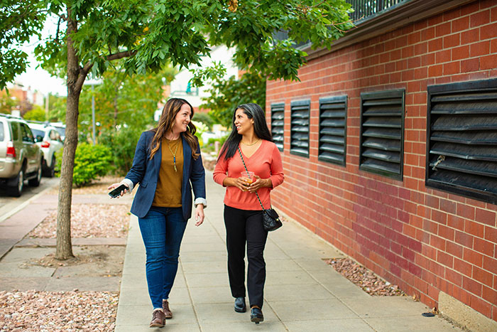 Two women walking and smiling on a sidewalk, sharing a touching conversation.