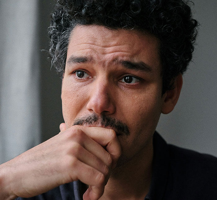 Man with curly hair and a mustache, looking thoughtful and touched, hand resting near his mouth.