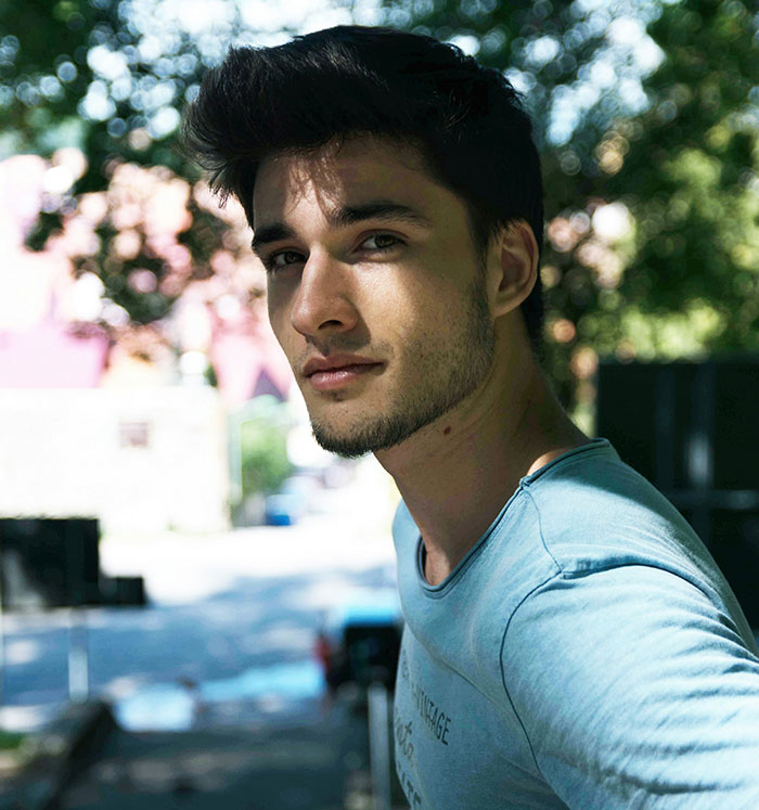 Young man in a blue shirt, outdoors, thoughtful expression, light filtering through trees, touching moment reflected in eyes.