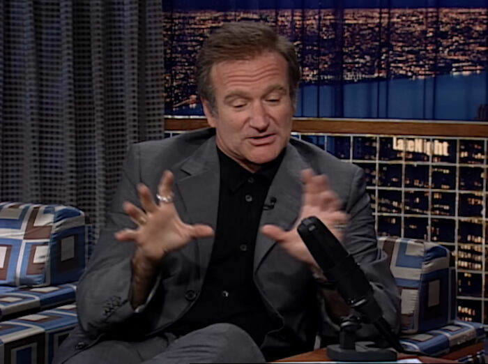 Man gesturing during an interview on a late-night talk show, with city lights visible in the background; celebrity deaths topic.