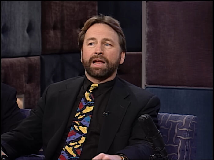 Man in a black suit with a colorful tie, seated on a patterned couch, discussing celebrity deaths.