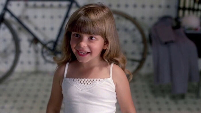 Young girl smiling, wearing a white dress, standing indoors.