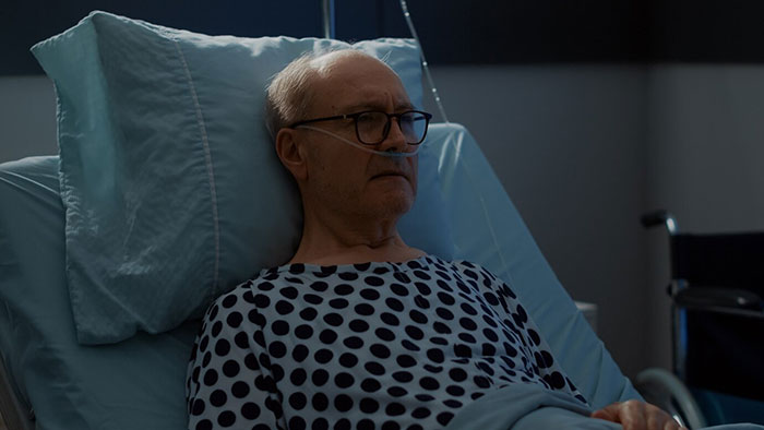 Elderly man in hospital gown, lying in a bed with medical equipment in the background.