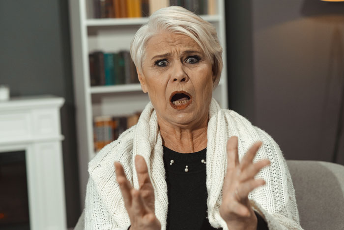 An elderly woman with a shocked expression, gesturing with her hands, sitting in a cozy room; focus on mother’s concern.