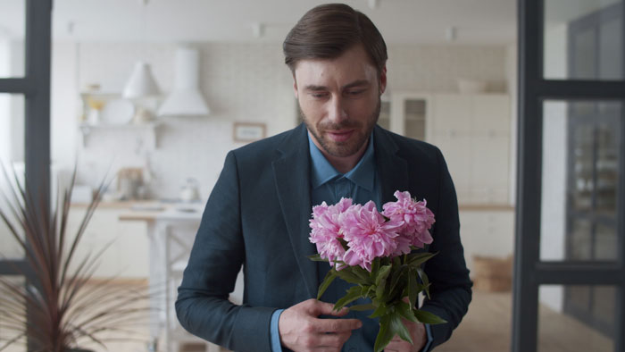 A man in a suit holding pink flowers in a modern kitchen, thinking about a family heirloom ring.