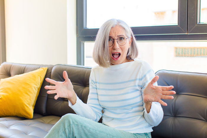 Elderly woman on sofa, looking surprised, wearing glasses and a striped sweater, gesturing with hands.