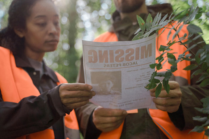 "Two people in safety vests examine a missing person flyer in a forest, highlighting man’s mystery disappearance.