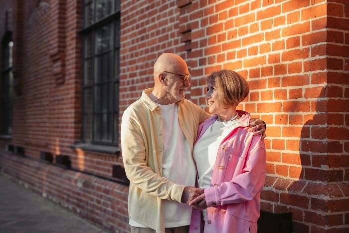 Elderly couple smiling and embracing outside, standing near a brick wall.