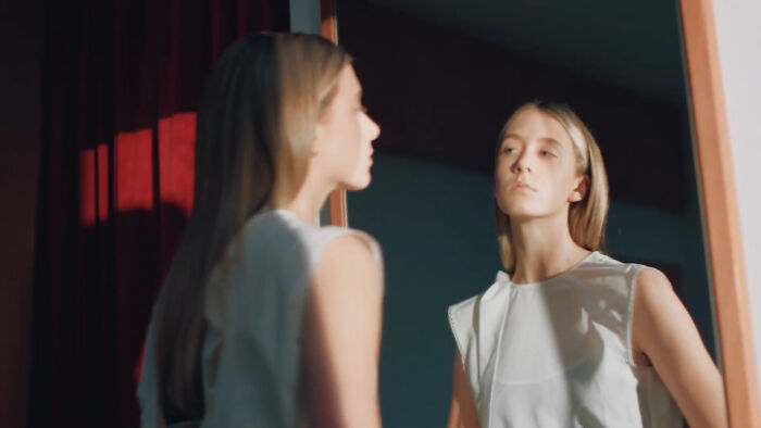 Young model gazing in mirror, wearing a white dress, in a softly lit room, embodying confidence and elegance.