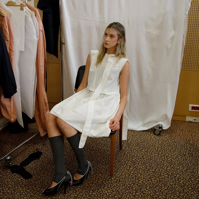 Young woman in white dress and black heels sitting, backstage at a fashion show, related to Nicole Kidman's "nepo baby.