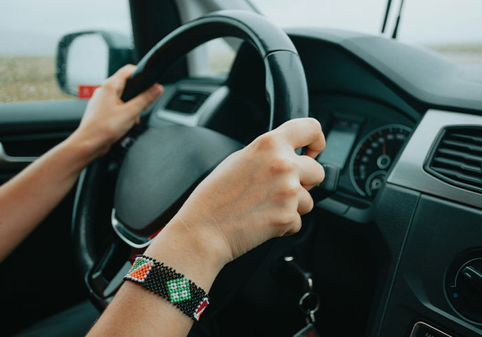 Hands on a steering wheel, wearing a colorful bracelet, inside a car; highlights normal aspects for US citizens.