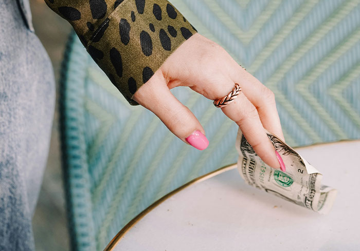 Hand with pink nails placing a US dollar bill on a table, representing normal US tipping culture.