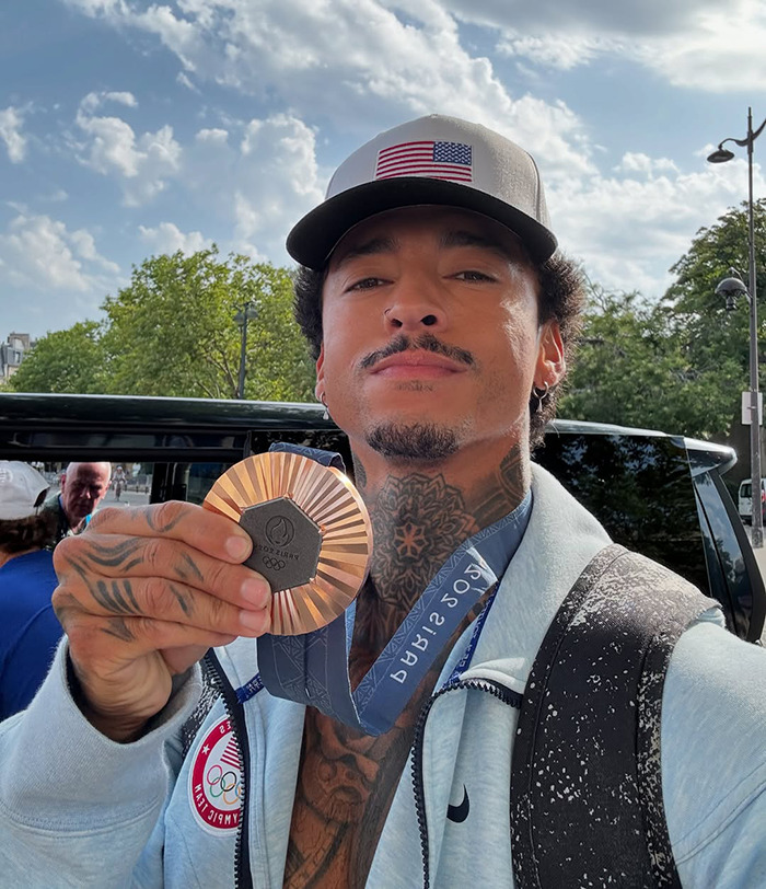 Athlete in Paris holding an Olympic medal outdoors, wearing a cap and light jacket.