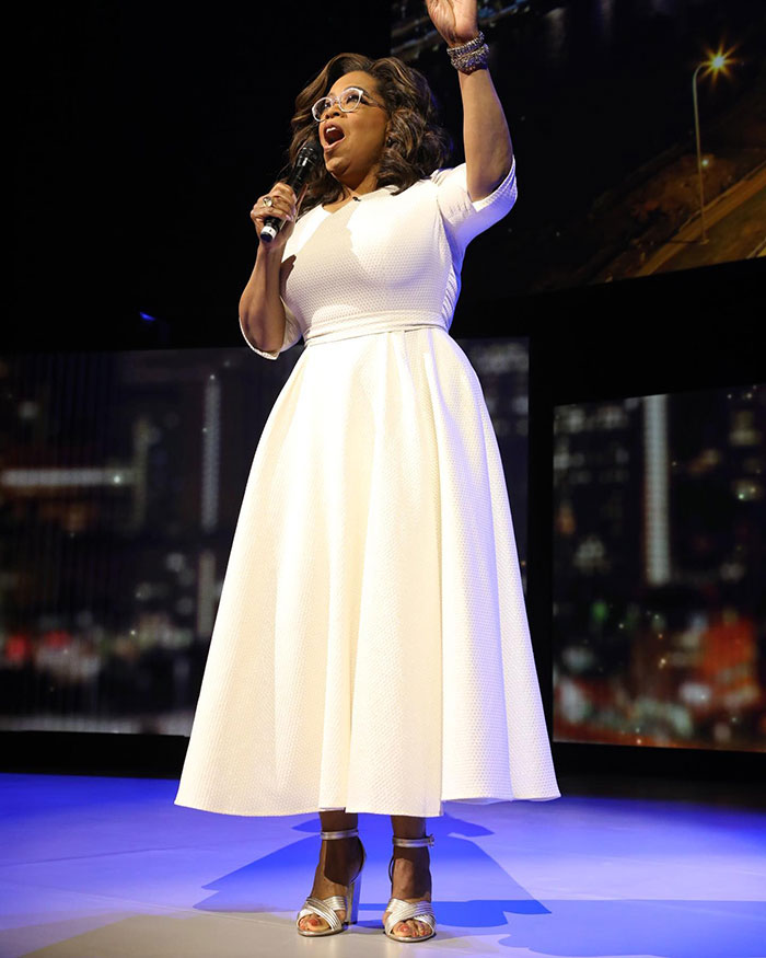 A woman on stage, wearing a white dress and holding a microphone, speaking passionately about thin people and Ozempic.