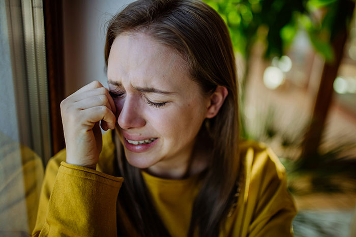 A woman in a yellow top, visibly upset and crying, sits by a window.