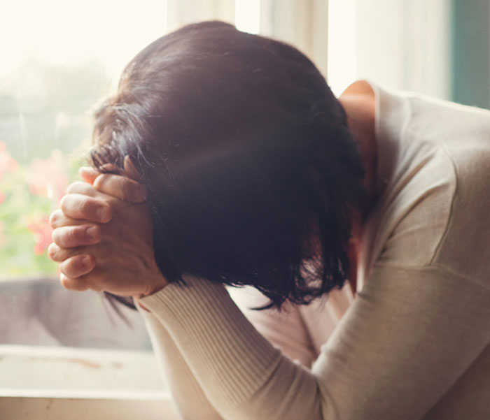 A woman sitting indoors, her head resting on clasped hands, appears to be in distress or deep thought.