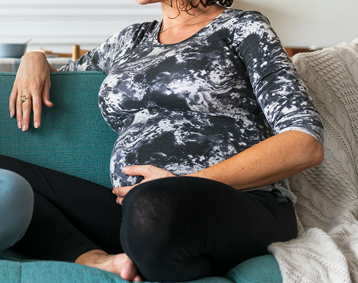 Pregnant woman sitting on a teal couch, wearing a patterned top, with hands on her belly.