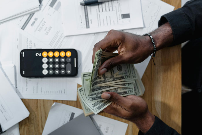 Man counting dollar bills near a smartphone calculator and mortgage documents on a table.