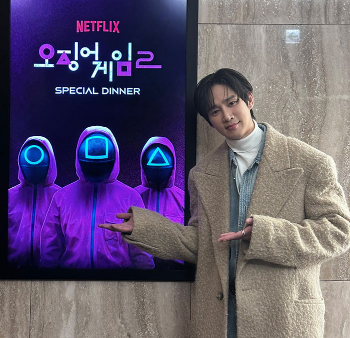 Actor with a poster for Squid Game special dinner event, wearing a beige coat and posing indoors.