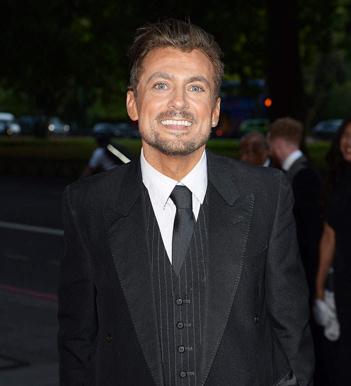 Love Island star in a black suit at an outdoor event, smiling at the camera.