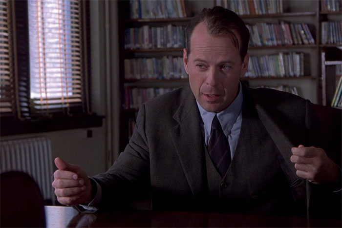 A person in a suit sitting at a table in a library setting, gesturing with their hands.