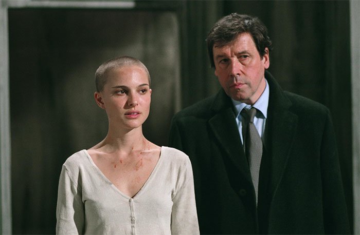 Two people standing in a dimly lit room; one with a shaved head, the other in a suit, conveying a serious atmosphere.