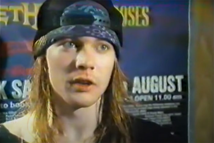 A person with long hair and a cap, standing in front of posters, unaware of their famous status.