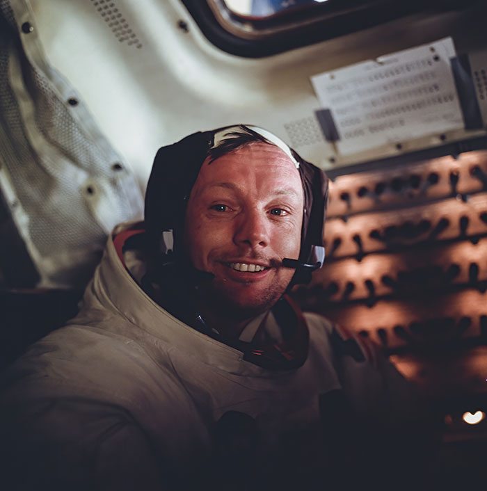 An astronaut smiling inside a spacecraft, wearing a space suit and helmet with communication gear.