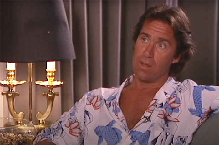 Man in a floral shirt sitting on a couch, beside a lamp with curtains in the background; theme of meeting famous people.