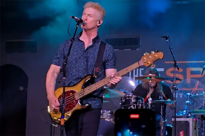 Musician performing on stage with a bass guitar, blue spotlight in background, person drumming behind.