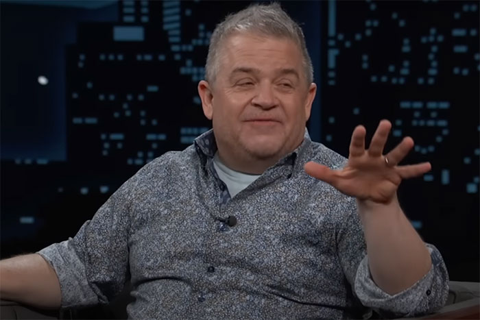 A person in a patterned shirt sits on a talk show set, gesturing with one hand.