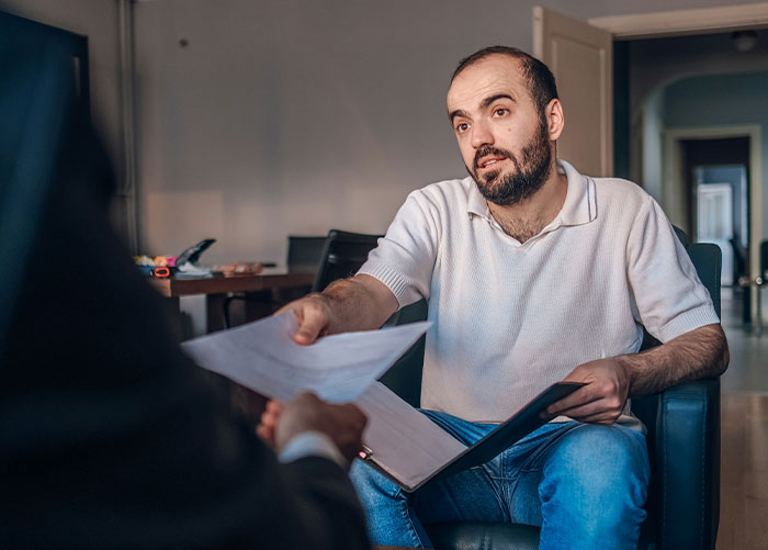 Man in a white shirt sitting and discussing while handing a document, evoking high school nostalgia.