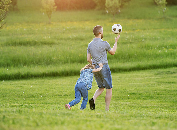 Man balancing a soccer ball while a child in jeans clings to his side, playing in a grassy field.