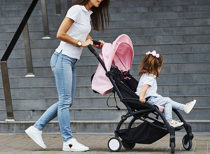 Woman pushing a child in a pink stroller, dressed casually, evoking nostalgia for high school peak behavior.